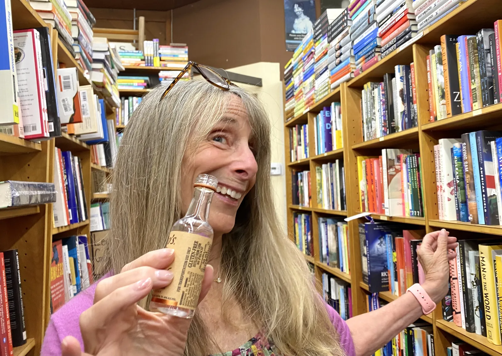 Sandy Pearl laughing, holding a miniature bottle among the shelves at Chaucer's Books